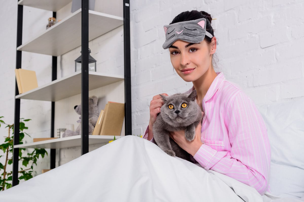 Woman wearing a cat-themed sleep mask holding her grey cat in bed.