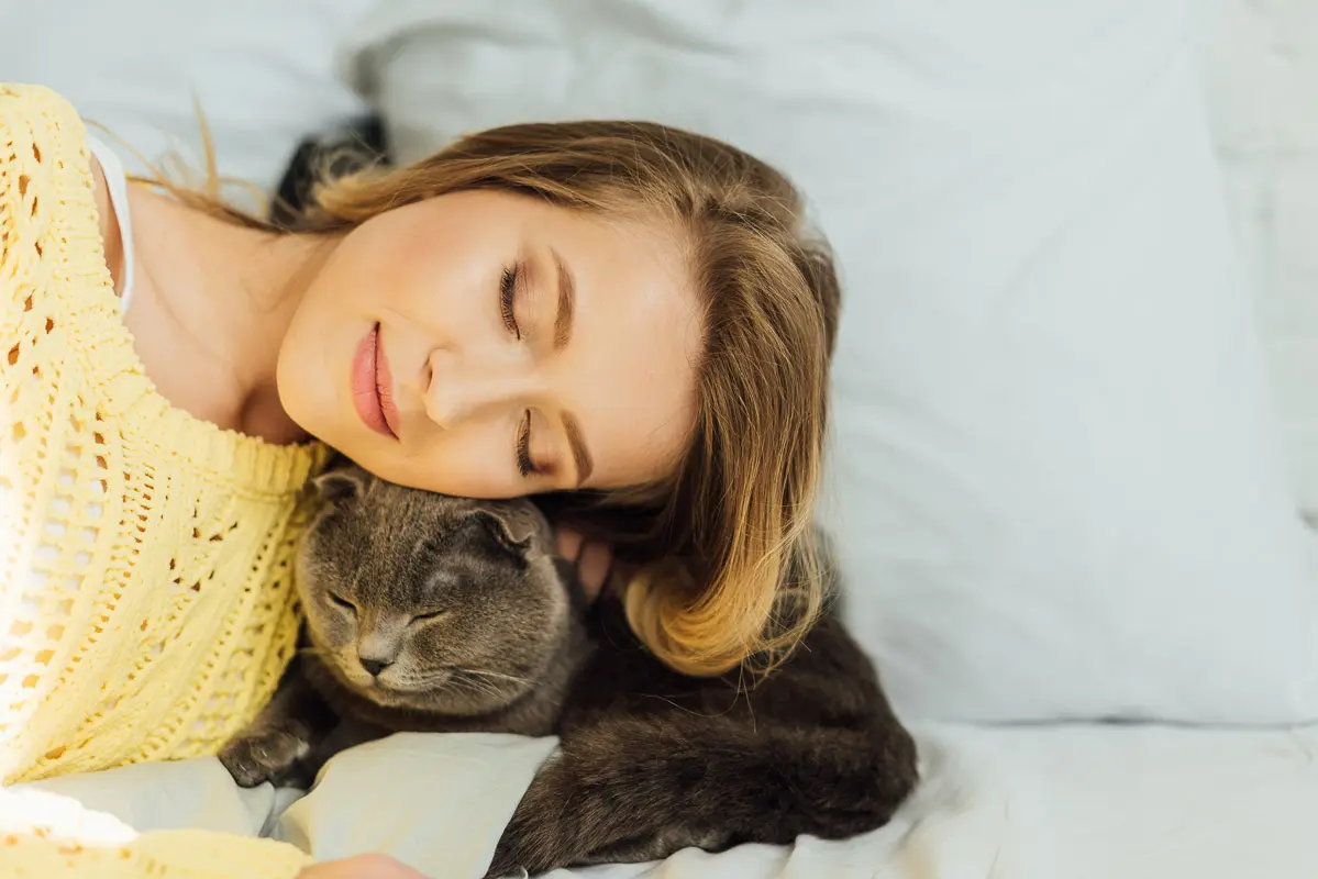 woman with eyes closed curled up with sleepy cat on bed